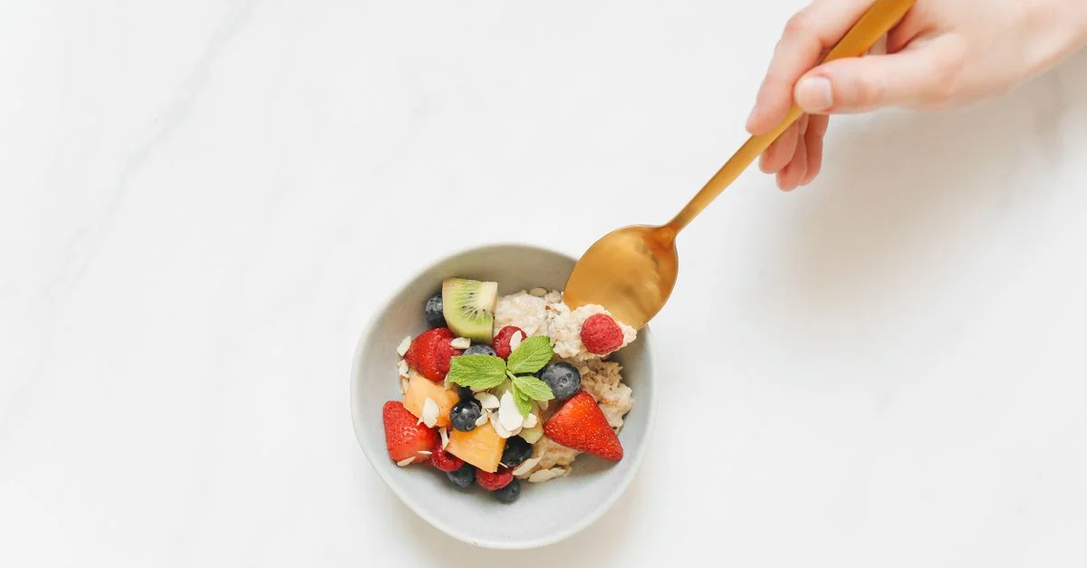 Vue de dessus d'un bol de petit-déjeuner coloré contenant des fruits et des flocons d'avoine, avec une main tenant une cuillère.