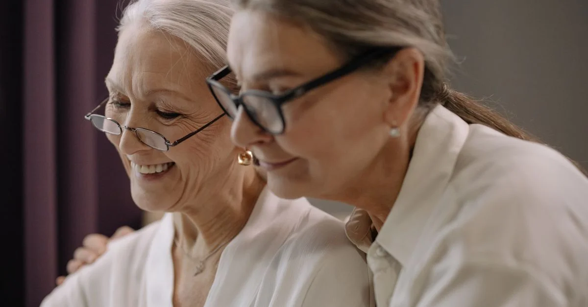 Deux femmes âgées portant des lunettes partagent un moment joyeux à l'intérieur.