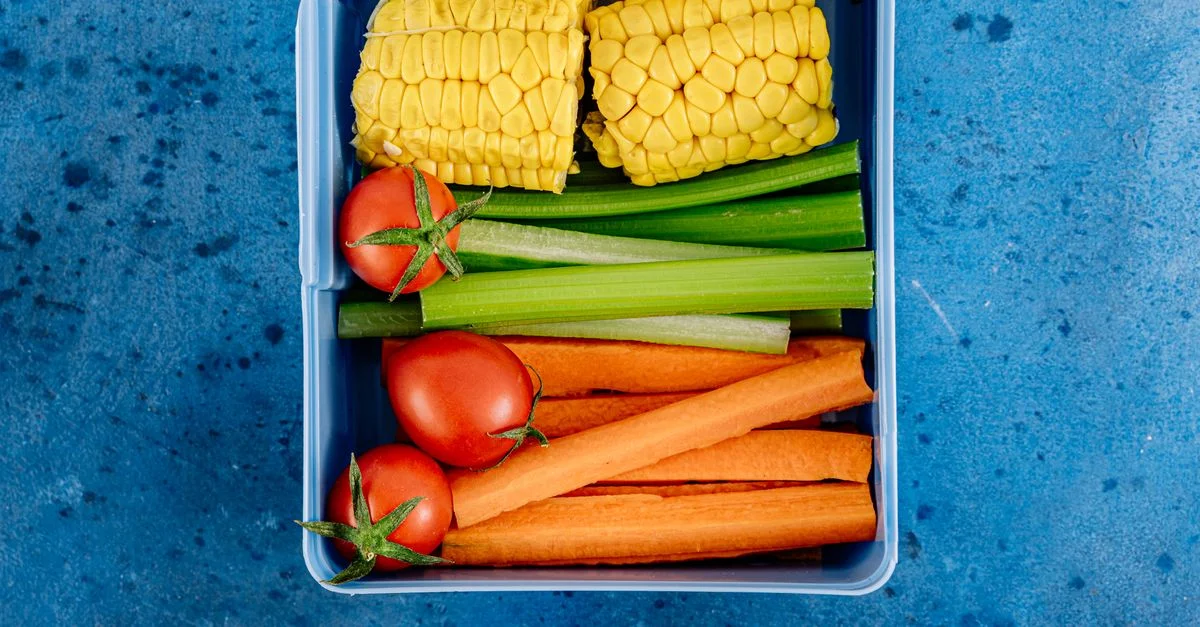 Vue de dessus d'une boîte à lunch remplie de légumes, maïs, tomates et collations sur un fond bleu.